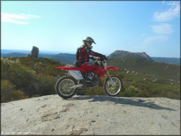 Scenic photo of man sitting on Honda CRF250X on top of solid rock ledge with rolling hills in the background.