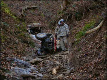 Two men wearing camouflage pants and jackets standing next to ATV that has turned over onto side on rocky trail.