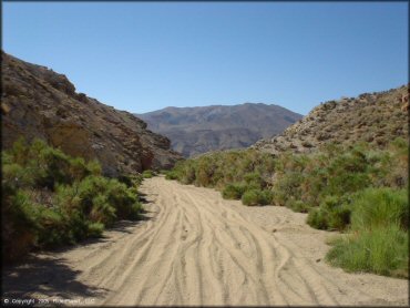 A section of a wide and sandy trail surrounded by rock ledges and creosote bushes.