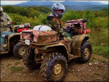 Woman wearing camouflage jacket and street helmet sitting on ATV with camping and hunting gear strapped to the front and back.