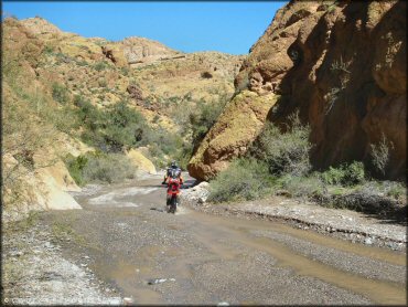 Honda dirt bike going through shallow stream with gravel and small river rocks.