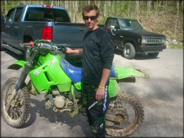 Man wearing Oneal camouflage motocross pants standing next to Kawasaki two-stroke parked behind blue GMC Sierra SL6.