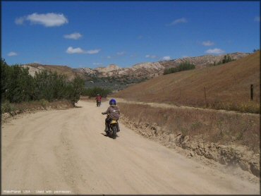 Two young children riding Suzuki 50cc dirt bikes on wide and flat 4x4 road.