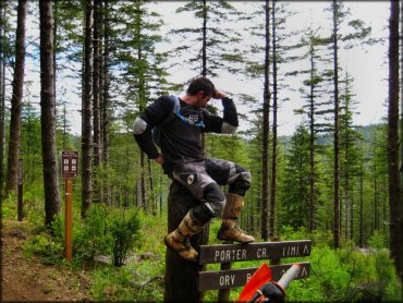 Man wearing Fox motocross jersey, CamelBak and Alpinestar motocross boots sitting on wooden stump off side of trail.