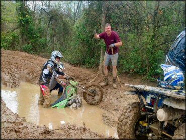 Three people using a tow rope pull Kawasaki two-stroke dirt bike from deep mud pit.