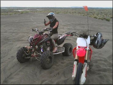 Young woman sitting on Yamaha Raptor 700R parked next to HOnda dirt bike with orange whip flag.