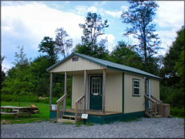 Cabin rental with front porch, two picnic tables, grassy backyard, stack of firewood and gravel parking.