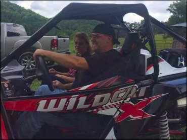 Arctic Cat Wildcat X with a man and woman sitting inside parked in staging area with antoher truck in the back.