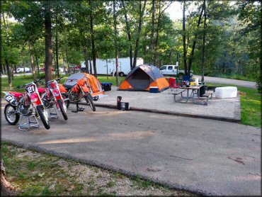 Campsite with three dirt bikes parked on concrete pad next to two tents, picnic table, lantern holder and BBQ grill.