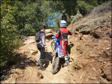 Three riders trying to get a Yamaha dirt bike up a rocky section.