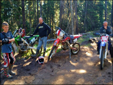 Young boy wearing Fox motocross pants, gloves and chest protector standing next to a Kawasaki, Honda and Yamaha dirt bike.