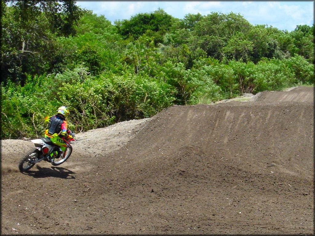 Man on dirt bike heading toward jump on motocross track.