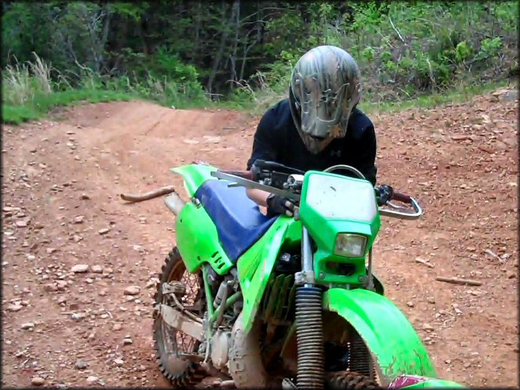 Man wearing camouflage motorcycle helmet standing next to Kawasaki KDX two-stroke with FMF Fatty Pipe on the trail.