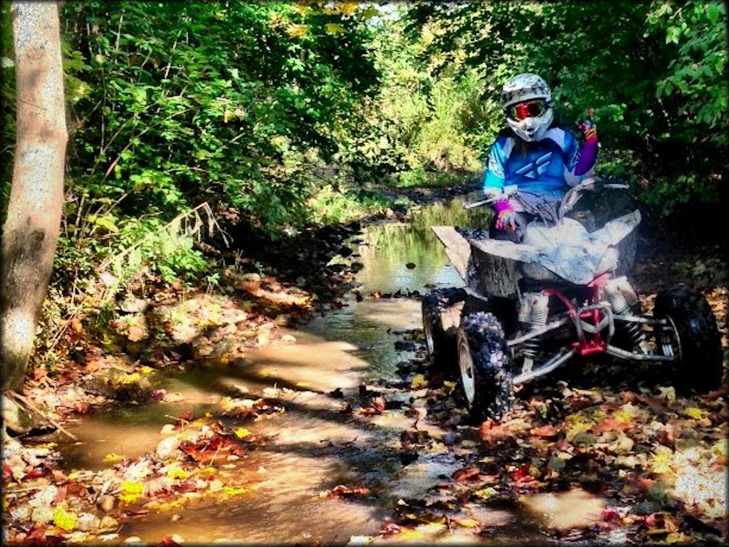 Rider wearing Fly motocross riding gear sitting on ATV with shallow creek in the background.