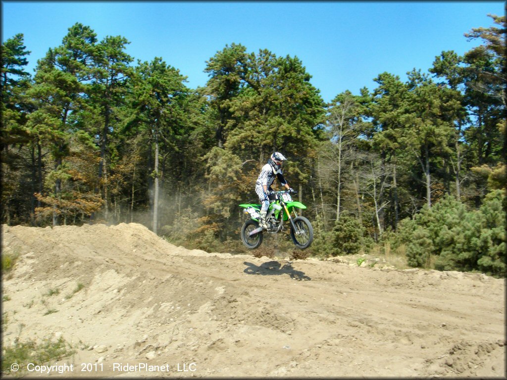 Kawasaki 4-stroke dirt bike going over jump on sandy motocross track.