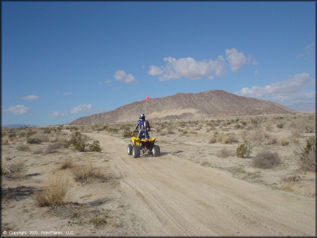 Woman on Yamaha Banshee 250 with red whip flag navigating wide sandy ATV trail.