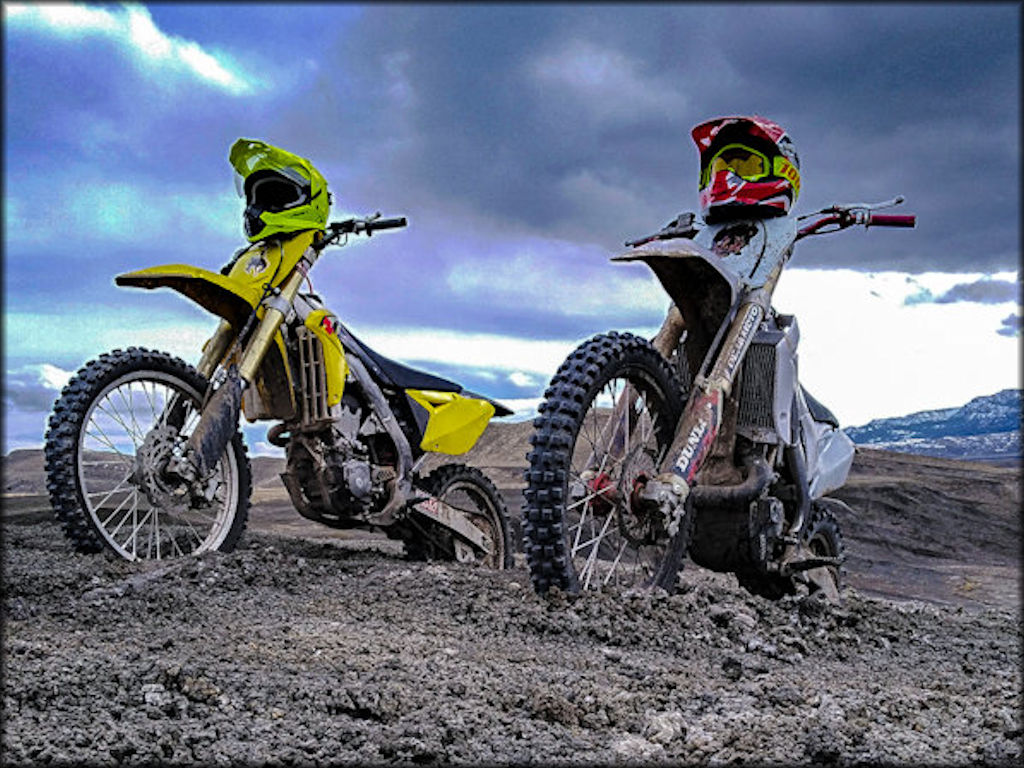 A Suzuki and Another 4-Stroke Motocross Bike with Helmets Propped on a Scenic Hilltop