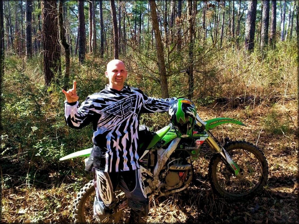 Man with MSR riding gear and green Kawasaki dirt bike parked off trail.