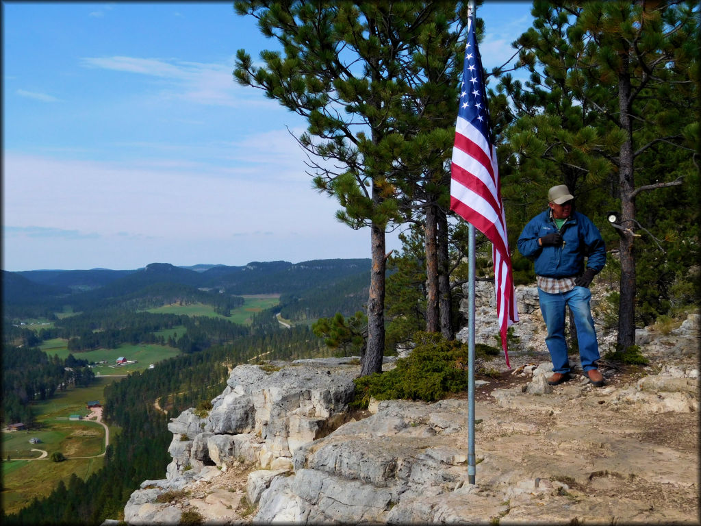 Elderly man standing next to flagpole with American flag overlooking cliff with scenic view of valleys, meadows and forests.