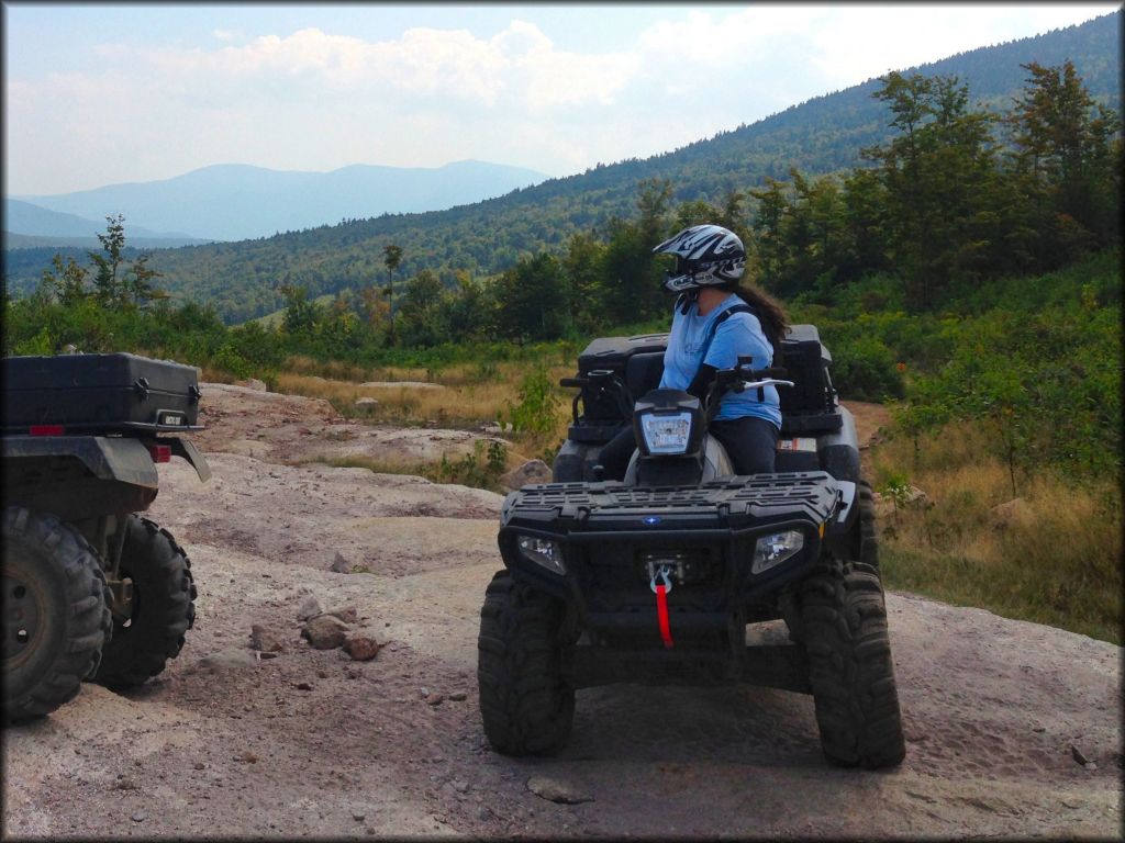 Woman sitting on Arctic Cat ATV with mud tires, rear cargo storage boxes and winch with red tow strap attached to front bumper.