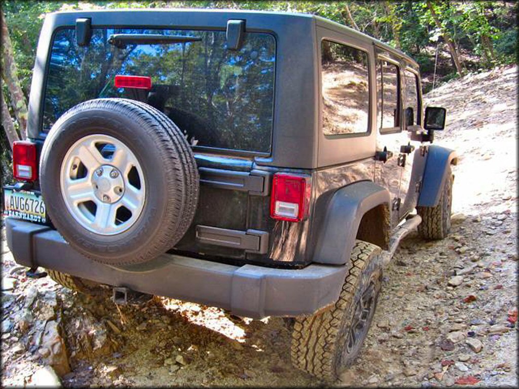 Hard top gray Jeep 4x4 with a GoodYear spare tire and custom rims on a rocky dirt road.