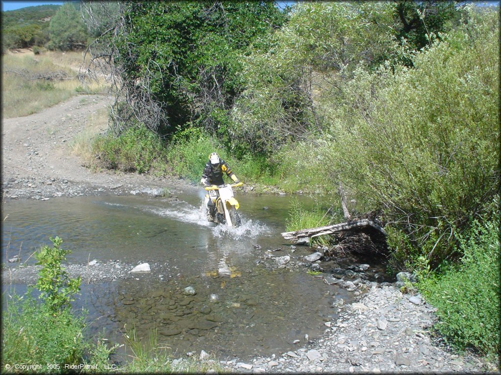 Man on RM250 dirt bike with yellow and black motocross gear riding through shallow creek crossing.