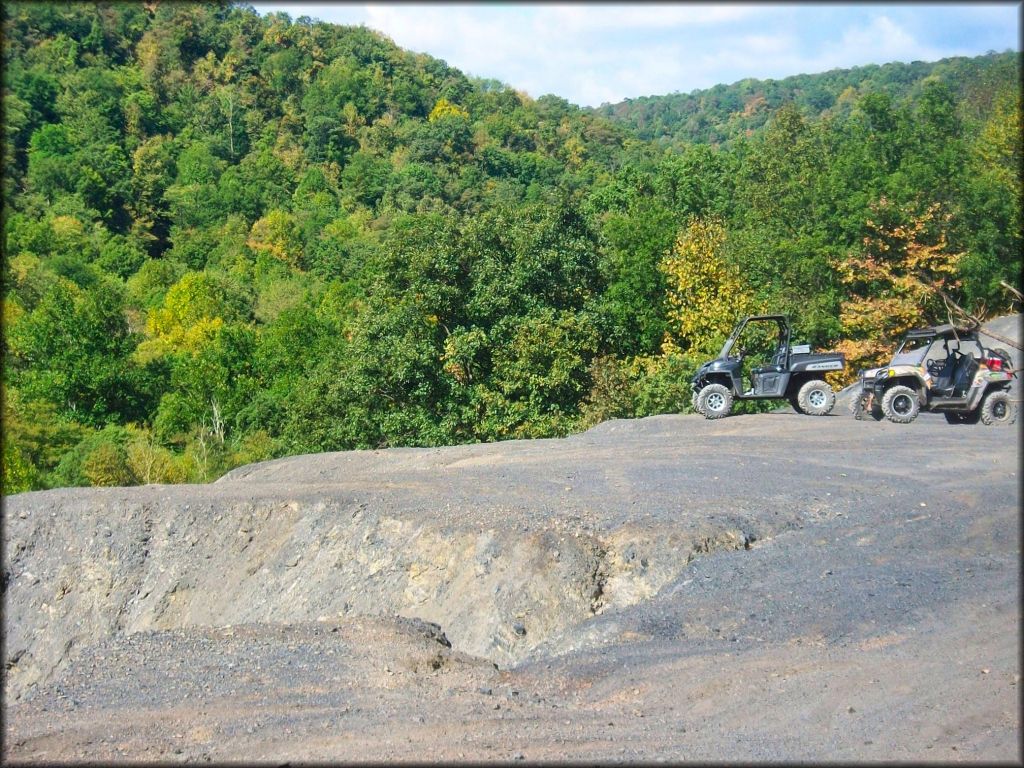 Two Polaris Ranger side by sides parked next to each other on top of gravel pit with trees in the background.