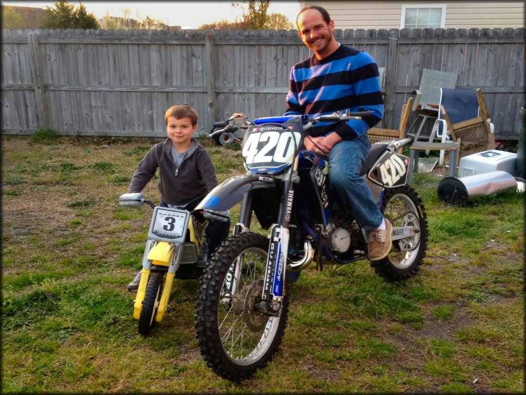 Father on black Yamaha YZ250 and young son on yellow mini dirt bike.