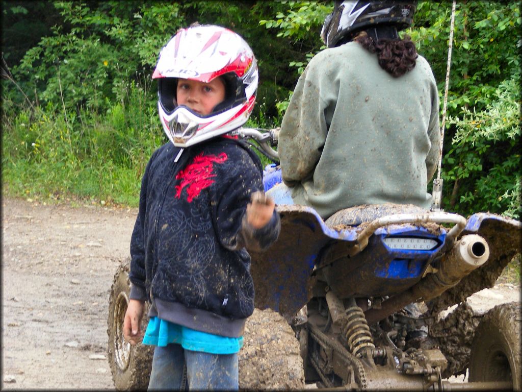 Young child wearing red and white motorcycle helmet standing next to woman sitting on Yamaha ATV covered in mud.