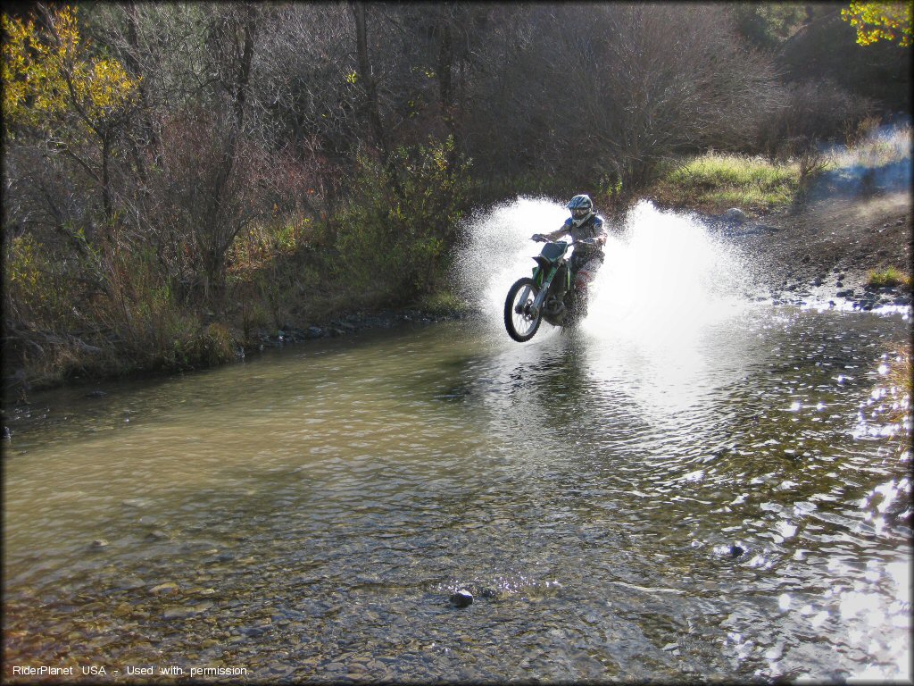 Kawasaki dirt bike doing a wheelie through shallow stream crossing.